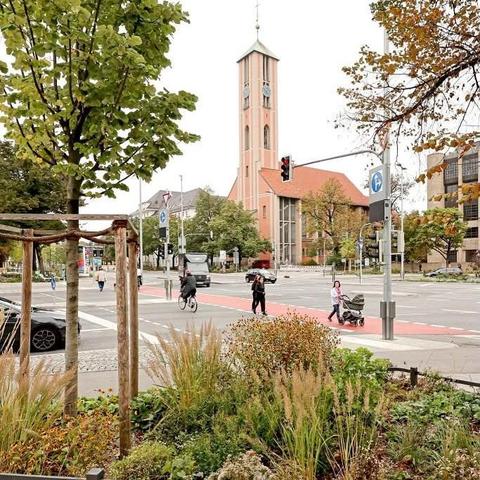 Das Bild zeigt eine Stadtstraße mit einem im Hintergrund stehenden Kirchturm, der eine auffällige rote Fassade und eine hohe, schlanke Turmspitze mit einem Zifferblatt aufweist. Vor dem Kirchturm befindet sich ein Kreuzungsbereich mit Fußgängern und Radfahrern. Einige Menschen gehen auf dem Bürgersteig, während ein Radfahrer die Straße überquert. Ein Kindwagen wird von einer Person getragen. Die Straße ist mit einem roten Radweg markiert, der sich durch die Szene schlängelt. Im Vordergrund sind gepflegte Pflanzen und Bäume zu sehen, die die Straße säumen. Die Umgebung wirkt urban, mit modernen Gebäuden im Hintergrund.
―