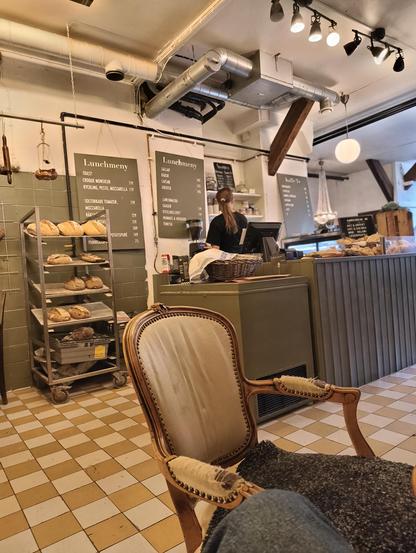 An interior shot of a cozy cafe or bakery. In the foreground is a large, ornate, antique wooden armchair with light-colored upholstery and a dark throw blanket. The background features a counter where a person with their back to the camera is working, likely taking orders or preparing food. Above the counter, two black chalkboards display the "Lunchmeny" (Lunch Menu) in Swedish, listing items like "Sallad," "Toast," "Caesar," "Croque Monsieur," and "Mozzarella." A metal shelving unit on the left holds several loaves of bread. The room has industrial exposed ductwork and a checkered yellow and brown tiled floor.