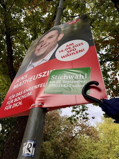 Wahlkampf der SPD Bochum-Ehrenfeld: Rotes SPD-Plakat von Jens Matheuszik mit zusätzlichem grünem Stichwahl-Sticker… der Schirm hilft beim Befestigen…