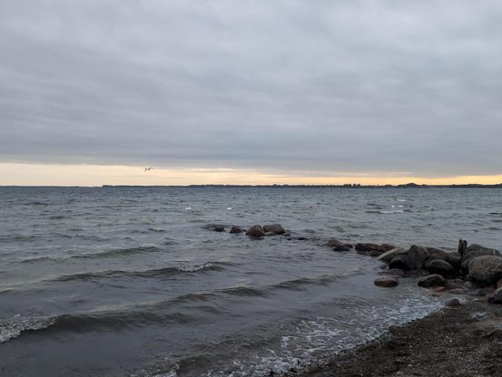 Ostseeblick. Wellige Wasseroberfläche. Eine Natursteinbuhne rechts im Bild. Grauer Himmel mit zart orange gefärbtem Streifen am Horizont.
