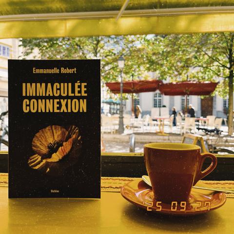 Photo de la couverture de l’ouvrage d’Emmanuelle Robert IMMACULÉE
CONNEXION avec à côté une tasse de café rouge. 
En arrière plan, la vitrine de botane, puis la place de l’hôtel de ville où on distingue deux parasols rouge, des tables, des chaises ainsi que la végétation des arbres