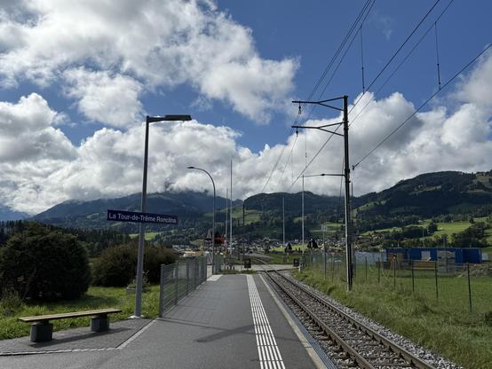 Train station platform with a sign for "La Tour-de-Trême Ronclina." The scene features nearby mountains, a clear blue sky with clouds, and railway tracks. A bench is present on the platform, and some greenery surrounds the area.