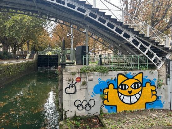 an aerosol-art piece of a cartoon cat anthropomorphically extending its arms as if offering a hug, on the side of a concrete bridge. To its left, a canal is visible