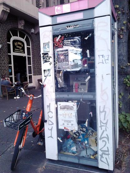 Foto einer völlig vermüllten, dreckigen, mit Graffiti beschmierten, mit längst obsoleten Plakaten beklebten Münztelefonzelle, an der ein Leihfahrrad (nur mit einem Smartphone benutzbar) gelehnt ist. Im Hintergrund sitzt jemand sehr bequem mit hochgelegten Beinen vor einem türkischen Restaurant und telefoniert mit seinem Handy.
