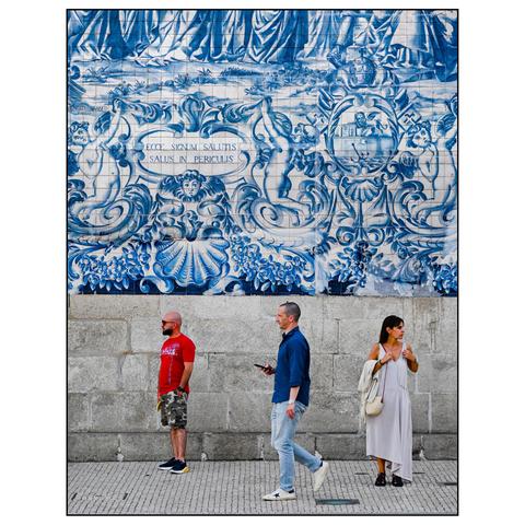 This image shows three people standing in front of a large, decorative tile mural. The mural features traditional Portuguese azulejos—blue and white ceramic tiles—depicting intricate, historical or mythological scenes with elaborate patterns, including figures, sea creatures, and ornate designs. The Latin phrase "ECCE SIGNUM SALUTIS SALUS IN PERICULIS" is prominently displayed on a banner within the mural, which translates to "Behold the sign of salvation, safety in danger."
The three individuals appear to be not noticing each other at all. One is standing looking to the left, the other is walking by watching his smartphone, the third is standing on the right, looking to the right. (draft by mistral.ai, edited by author)