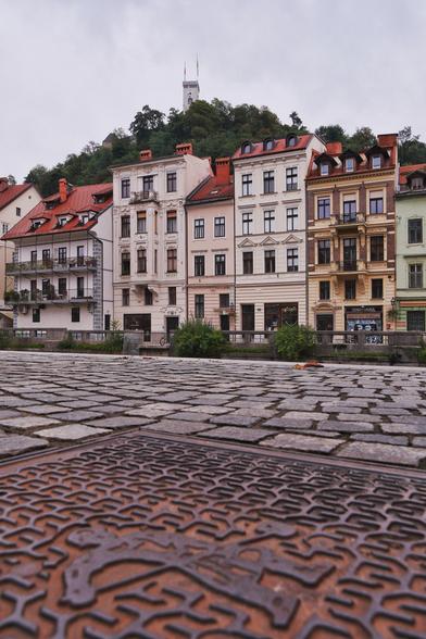 Häuser der Altstadt unterhalb der Burg in Ljubljana. Die Häuser stehen entlang der Ljubljanica. Im Vordergrund Boden mit Kanaldeckel.