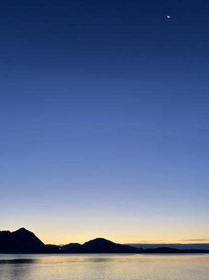 A dawn scene featuring a calm lake reflecting the sky. Silhouetted mountains are visible in the foreground against a gradient sky transitioning from deep blue to warm orange near the horizon. A crescent moon is visible in the upper right corner.