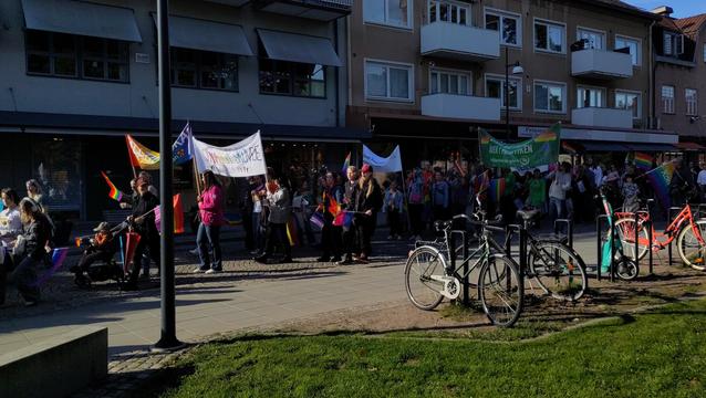 People on the sunlit streets of downtown Skövde in Sweden carrying banners and pride flags.