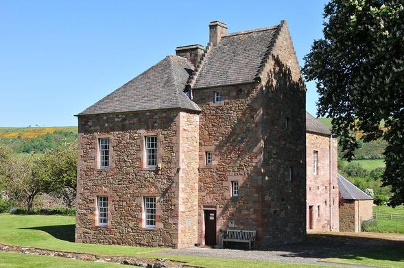 The Commendator's House Museum. The image shows a large pink and honey-coloured stone building with grey slate roofs. It is two storeys in height apart from a three-storey tower on the right side of the end nearest us. The building is on a slope leading away from us down into a broad shallow valley and there are fields and trees visible on the far side. There’s a tree on the right side of the frame and the sky is blue.