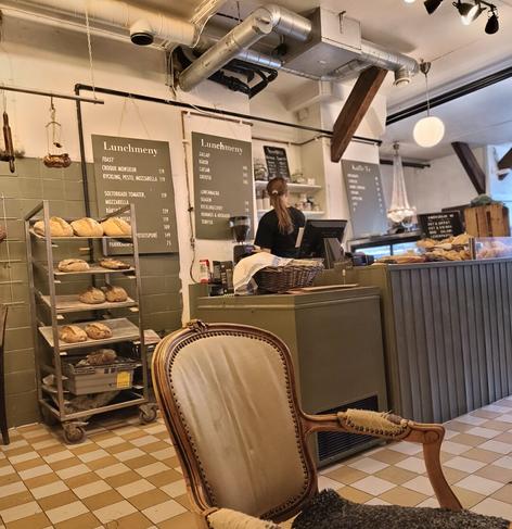 An interior shot of a cozy cafe or bakery. In the foreground is a large, ornate, antique wooden armchair with light-colored upholstery and a dark throw blanket. The background features a counter where a person with their back to the camera is working, likely taking orders or preparing food. Above the counter, two black chalkboards display the "Lunchmeny" (Lunch Menu) in Swedish, listing items like "Sallad," "Toast," "Caesar," "Croque Monsieur," and "Mozzarella." A metal shelving unit on the left holds several loaves of bread. The room has industrial exposed ductwork and a checkered yellow and brown tiled floor.