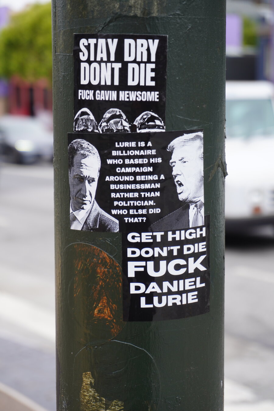 Several polical stickers on a post. One shows a photo of Daniel Lurie and Donald Trump with text that says "Lurie is a billionaire who based his campaign around being a businessman rather than politician. Who else did that?"  Another sticker says "Get high, Don't Die, Fuck Daniel Lurie."  The stickers are white text on black background and all stuck on a street sign post on Valencia St in San Francisco.
