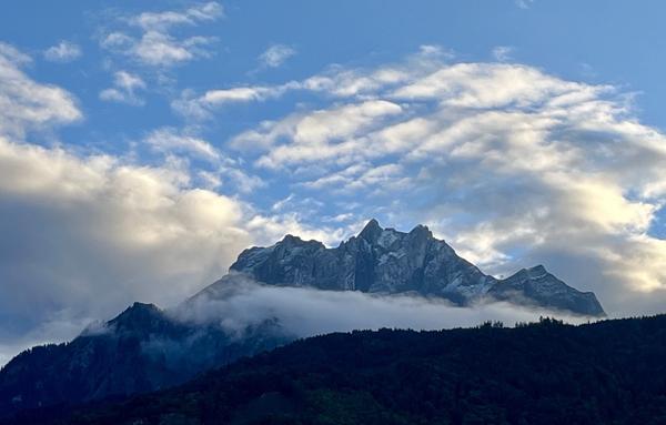 Pilatusberg, ganz leicht mit Schnee bestäubt, nach Sonnenuntergang. Ein Nebelband zieht vor dem Berg durch. Der Himmel ist blau, mit zahlreichen weiss-graue Wolken, die teilweise noch von der Sonne beschienen werden. Die bewaldeten Hügel vor dem Berg erscheinen fast schwarz.