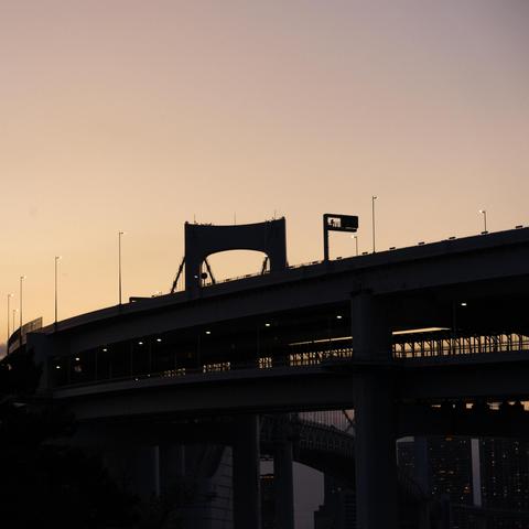 Silhouette of a highway bridge during twilight, with streetlights evenly spaced along its length. The sky is a gradient of warm colors, transitioning from light orange to deep purple. Bridge arch and support beams are visible against the sky. Dark outline of trees or foliage in the lower left corner.