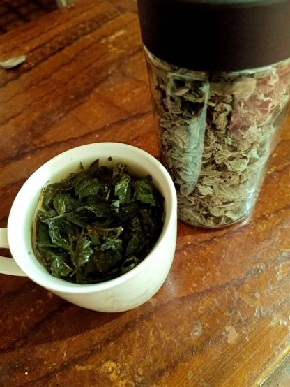 A teacup with green leaves floating around in some yellow-green water. Next to it a glass jar with black plastic lid stuffed full of dried, greenish leaves.