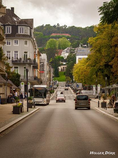 Blick entlang der Parkstraße in Bad Nauheim: Eine belebte Straße mit vorbeifahrendem Bus, Autos und Passanten, eingerahmt von eleganten Häusern und herbstlich gefärbten Bäumen. Im Hintergrund erhebt sich ein bewaldeter Hügel. Eine Szene, in der sich städtischer Alltag und Natur harmonisch begegnen.
