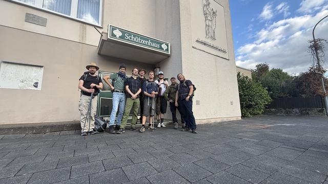 A group of seven individuals poses in front of a building labeled "Schützenhaus." They are dressed in casual outdoor clothing, equipped with backpacks and walking sticks. The setting features a paved area with clouds visible in the sky.
