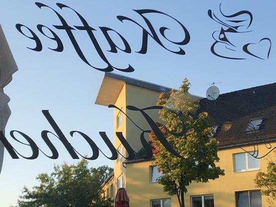 Durch Fenster fotografiert das beschriftet ist mit „Kaffee…“. Draußen ein leuchtend gelbes Haus gegenüber, blauer Himmel, ein junger Baum, die Blätter an einer Seite verfärbt.
