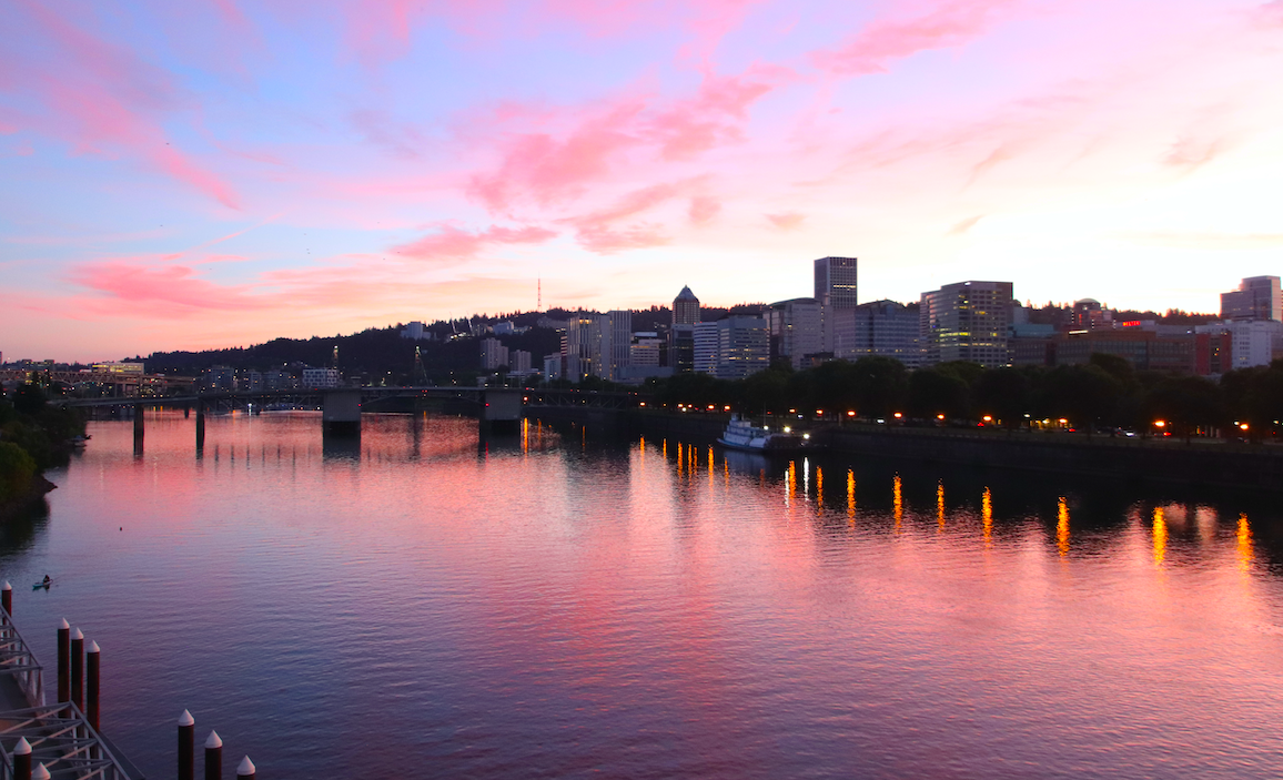 a wide river reflects the pink and pastel blue sunset. on the right, the lights and structures of a small city are seen. a boat floats leisurely down river, and a bridge crosses it in the distance