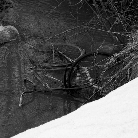 Black and white photo of a bicycle submerged in a shallow stream in winter, surrounded by branches, with the embankment covered in snow.