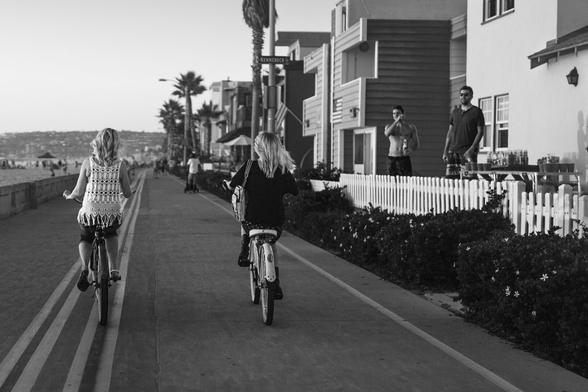 Black and white photo of two women riding bicycles down a beachside boardwalk, their backs to the camera. One wears a crocheted top, the other a dark sweater with a bag slung over her shoulder. Palm trees, beach houses, and the ocean stretch along the sides, with two men standing behind a white picket fence:;one shirtless, holding a drink, the other in sunglasses. The men may be saying something to the women as they pass.