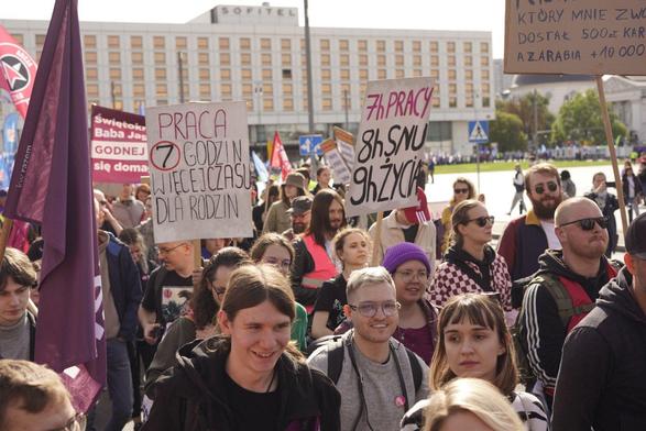 Tłum maszerujących ludzi. Widać flagę  Razem oraz transparenty "Praca 7 godzin więcej czasu dla rodzin" oraz "7h pracy 8h snu 9h życia".
