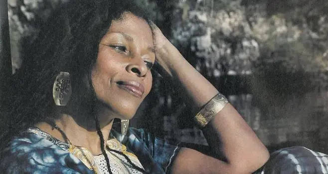 A photo of revolutionary sister Assata Shakur as a younger woman, with brown skin and long locs wearing large metal earrings and a blue dashiki with yellow and white embroidery. She has a soft smile on her face with her head resting against her hand.