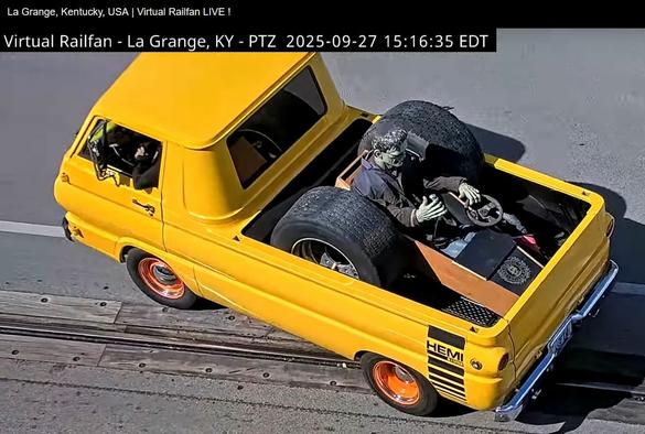 Ein alter gelber Minitruck mit einem nachgebautem Drag-Race-Fahrzeug auf der Ladefläche, in welchem eine Frankensteinfigur sitzt - Blick von oben

Das Fahrzeug steht in La Grange - Kentucky - USA auf einer Straße mit einer Schiene, auf welcher sporadisch lange Güterzüge fahren.
Dort findet ab und zu ein Treffen aufgearbeiteter alter Fahrzeuge statt