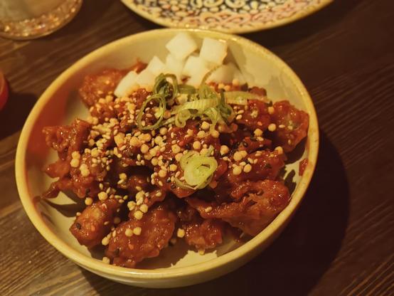 A close-up, top-down shot of a light-colored ceramic bowl filled with pieces of reddish-brown Korean fried oumph coated in a glistening, thick, spicy-sweet yangnyeom sauce. The dish is heavily garnished with small, light-colored sesame seeds and what appear to be crushed peanuts or other crunchy cereal/nut pieces. Sliced green onions are sprinkled over the top. The oumph is served alongside a small mound of white pickled daikon radish cubes at the top of the bowl. The bowl sits on a dark wooden table, with another blurred dish visible in the background.