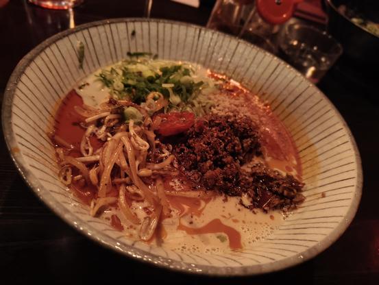 A close-up shot of a dark-rimmed, vertically striped ceramic bowl containing a dish of noodles in a creamy, rich broth with a significant layer of spicy red chili oil visible on the surface. The dish is topped with several distinct ingredients arranged in sections: a large mound of dark, coarsely ground seasoned vegan beef; a pile of reddish-brown bean sprouts; a scattering of finely chopped green onions and other white vegetables; and a slice of cherry tomato. The broth shows a creamy base (from coconut milk) mixed with the red chili oil, creating an appealing, marbled pattern. The bowl is set on a dark, indistinct surface, with blurry restaurant ambiance and glassware visible in the dark background.