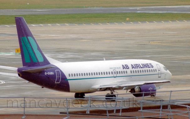 Side view of a twin engined jet airliner taxiing from left to right.
The plane is mostly white, with purple "AB Airlines" on the upper forward fuselage, with smaller green "First For Value" text aft of that.
A thin green stripe runs along the body, under the passenger cabin windows.
The rear fuselage and tail is purple, with the white registration on top of the fuselage, and large, green, letters "AB" on the tail.
The roof of a terminal building fills the foreground, with metal railings on top .
Grey concrete apron fills the background, with green grass lining the sides of a black runway in the distance.