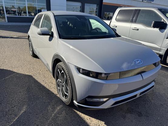 The image features a silver Hyundai electric vehicle parked in a lot. The car has a sleek, modern design with a smooth, aerodynamic body and distinctive LED headlights. The front grille is closed, a common feature in electric vehicles, and the Hyundai logo is prominently displayed in the center. The car is equipped with stylish alloy wheels and has a compact, hatchback shape. The background shows a building with large windows and another vehicle, possibly a white SUV, parked next to it. The setting appears to be a dealership or a car sales lot, with a clear sky and bright sunlight casting shadows on the ground.
