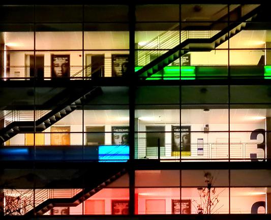 A modern, glass-fronted building lit up at night with different colours for different floors.