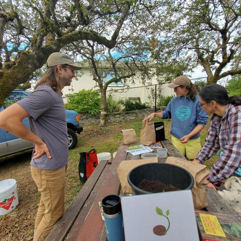 The image shows three people gathered around a weathered wooden picnic table, participating in a seed-planting workshop outdoors under a large tree. A man with long hair and a baseball cap stands to the left, facing the table with his hands on his hips. Two women are standing on the right side near the table. The first women is wearing a blue hoodie , and yellow short facing the other woman that is in a red/white/blue plaid shirt. There's a blue van visible in the background, parked on a grassy area. The table is covered with various seed packets, brown paper bags, a bucket filled with soil, and gardening supplies such as small containers and a book titled "Plants of the Pacific Northwest Coast". A white board with a drawing of a seed with a seedling sticking out of it is placed on the table, and a label of "Seed Planting Event" is attached to the bucket. The ground is covered with fallen leaves