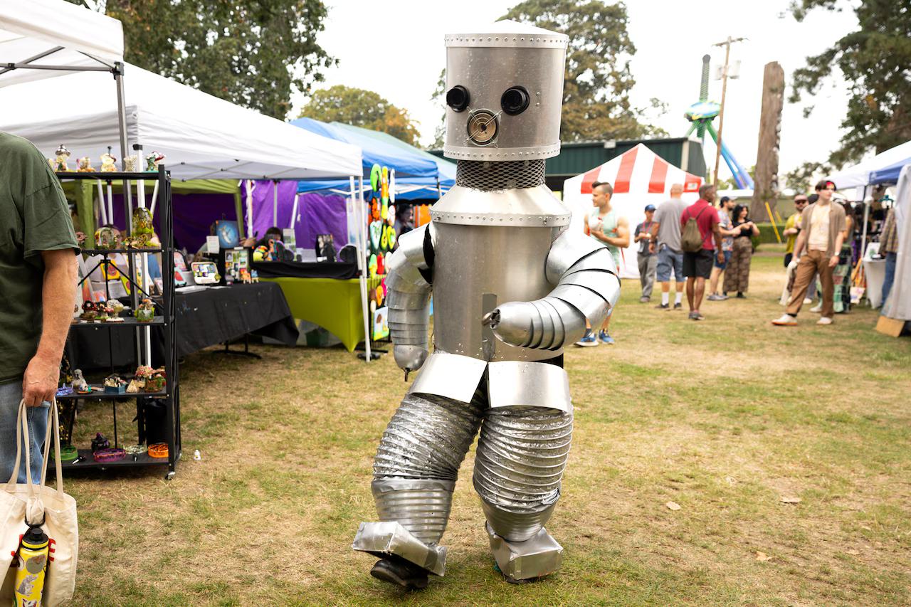 a person in a fantastic silver robot costume walking through a festival with various vendor stalls.