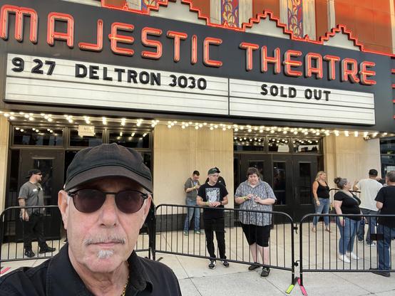 This photo shows the front of the historic Majestic Theatre, with its distinctive red neon signage prominently displayed. The marquee reads “927 DELTRON 3030” on the left side and “SOLD OUT” on the right, indicating a popular concert that has reached capacity.

In the foreground, I am taking a selfie - I’m wearing dark sunglasses, a black cap, and have a gray mustache and goatee. I am dressed in black clothing.

Behind me, you can see a crowd of people waiting outside the theater, contained by metal barriers. The theater’s facade features classic architecture with decorative elements, and the entrance is illuminated by strings of small lights creating a festive atmosphere. The building has an ornate, vintage movie theater design typical of early-to-mid 20th century entertainment venues.

The scene captures the energy and excitement of a sold-out show, with concertgoers gathered outside this iconic venue. The lighting suggests this photo was taken in the evening, just before or during the event.​​​​​​​​​​​​​​​​