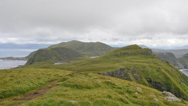 A photo of the green hilly landscape of a large island. There is a trail visible, and several small lakes on the island. The sky is cloudy.