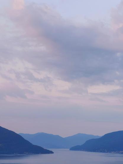 A tranquil lake bordered on the right and left by mountains bathed in the fading shadows of night. The mountains in the foreground show a deeper blue than those in the background. The sky is a delicate blue and pink, with light clouds shimmering in pastel shades ranging from blue to pink to violet. The image conveys an atmosphere of peace and transcendence.