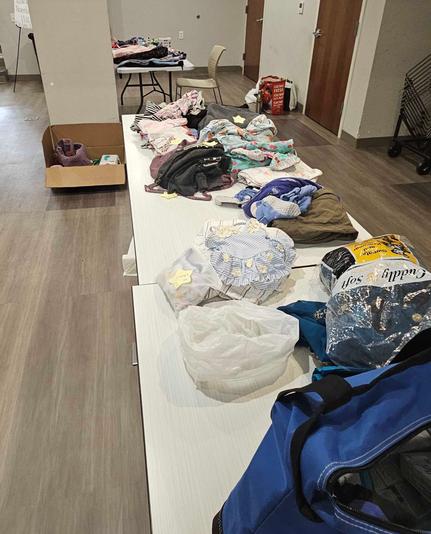A photo of two long white plastic tables in a row with clothing and other items on them, and some boxes and other items in the background in a large room.