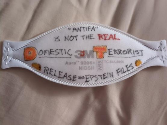 A 3M Aura mask (respirator) with the phrases ""Antifa" is not the REAL Domestic Terrorist" and "Release the Epstein Files" written on it in marker. The D and T are enlarged and coloured orange, with a small orange J on the 3M logo in the center. The bottom phrase is bookended by taco emoji.