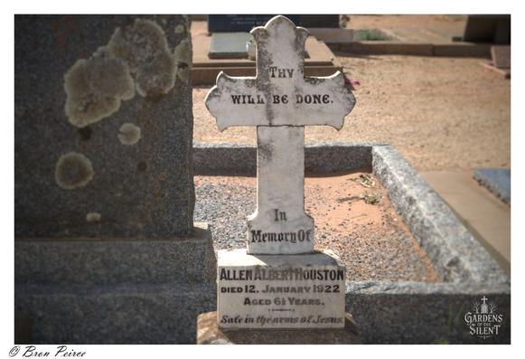 Stone cross shaped gravestone for Allen Albert Houston, aged 6½ years, inscribed with “Thy will be done” and “Safe in the arms of Jesus.”  Set on a weathered base amid other markers and a dirt path, framed by quiet reverence.  Watermarked “Gardens of the Silent” with signature by Bron Peirce.