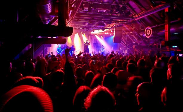 Totale der Bühne, kräftiges rotes und oranges Licht. Die ganze Band ist sichtbar, der Sänger steht erhöht, das Publikum feiert dicht gedrängt, Hände in der Luft, Club-Atmosphäre mit Dachgeschoss-Decke und Balkenkonstruktion.
