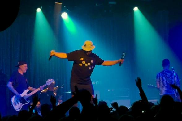 Nahaufnahme der Bühne. Der Sänger steht in der Mitte, Arme ausgebreitet, Mikrofon in der Hand, trägt weißen Fischerhut und schwarzes T-Shirt. Gitarrist und Bassist links, buntes Bühnenlicht, das Publikum jubelt mit erhobenen Händen.