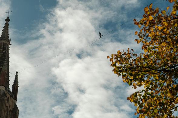Tightrope walker balancing above Sashuisbrug in Brugge against a cloudy sky.