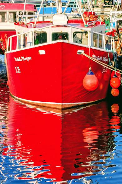 A close-up, vertical shot of the bright red bow of the fishing trawler docked in a harbour. The boat's vibrant red hull is intensely reflected in the calm, deep blue water below, creating a striking contrast and a shimmering, inverted image. Two small, bright orange buoys hang from the boat's side, mirrored in the reflection.