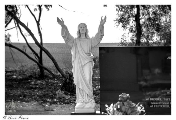 Black and white cemetery photograph featuring a statue of Jesus Christ with raised arms, robed and serene, standing before a dark polished gravestone.  Trees, fencing, and open field form a tranquil backdrop.  Watermarked “Gardens of the Silent” with signature ©Bron Peirce.