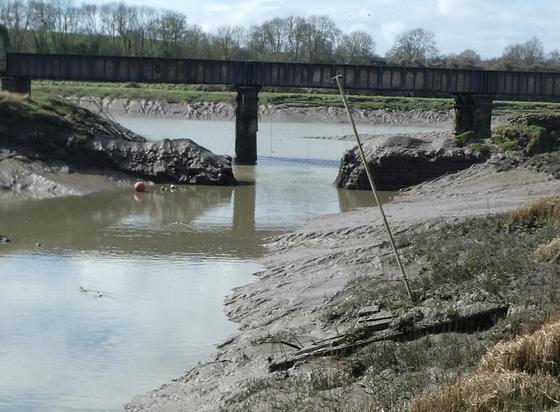 A muddy stream, meeting a larger river, under a bridge. In the foreground, an abandoned sailboat.