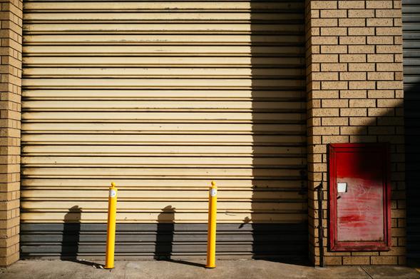A corrugated, cream-coloured garage door is closed, but the morning sun shines upon it, casting shadows from two bright yellow bollards in front of it. To the right is a fire hose door.