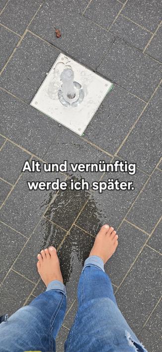 Zwei, vin oben aufgenommene Barfüße am Ende einer Jeans stehen in einem flachen Brunnen. Über den Füßen sprudelt ein kleine Fontaine. Der Boden des Brunnens besteht aus verschiedenen großen rechteckigen Pflastersteinen.