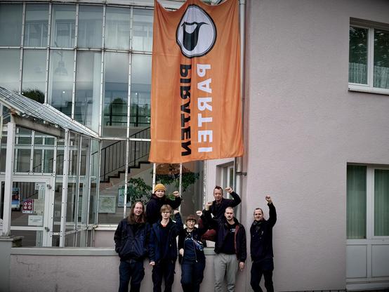 7 Personen mit hochgestreckten Fäusten vor einem Gebäude, an dem eine große orange Flagge der Piratenpartei hängt.