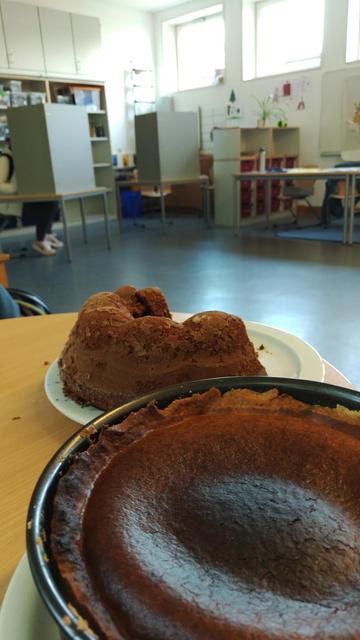 Zu sehen sind im Vordergrund zwei Kuchen, im Hintergrund zwei Wahlkabinen. In einer Wahlkabine sitzt jemand, die Beine sind zu sehen.